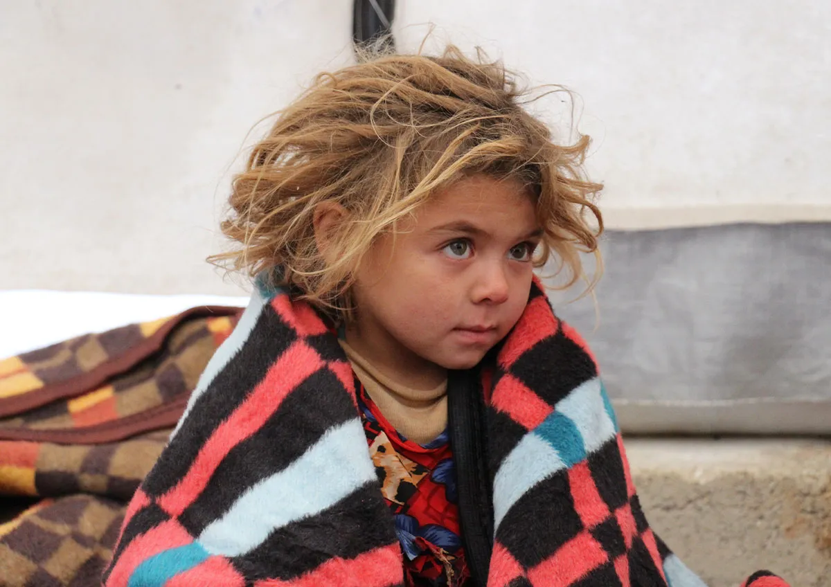 Un niño pequeño afectado por la crisis de los refugiados sirios está sentado en una tienda de campaña con una manta a rayas envuelta alrededor. Los refugiados en Siria luchan por acceder a necesidades básicas como comida, refugio y atención médica.