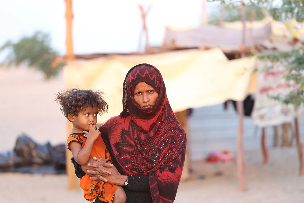 A woman wearing a red patterned hijab holds her young child on her hip. Her child is wearing a bright orange garment. Behind them is a white makeshift tent.