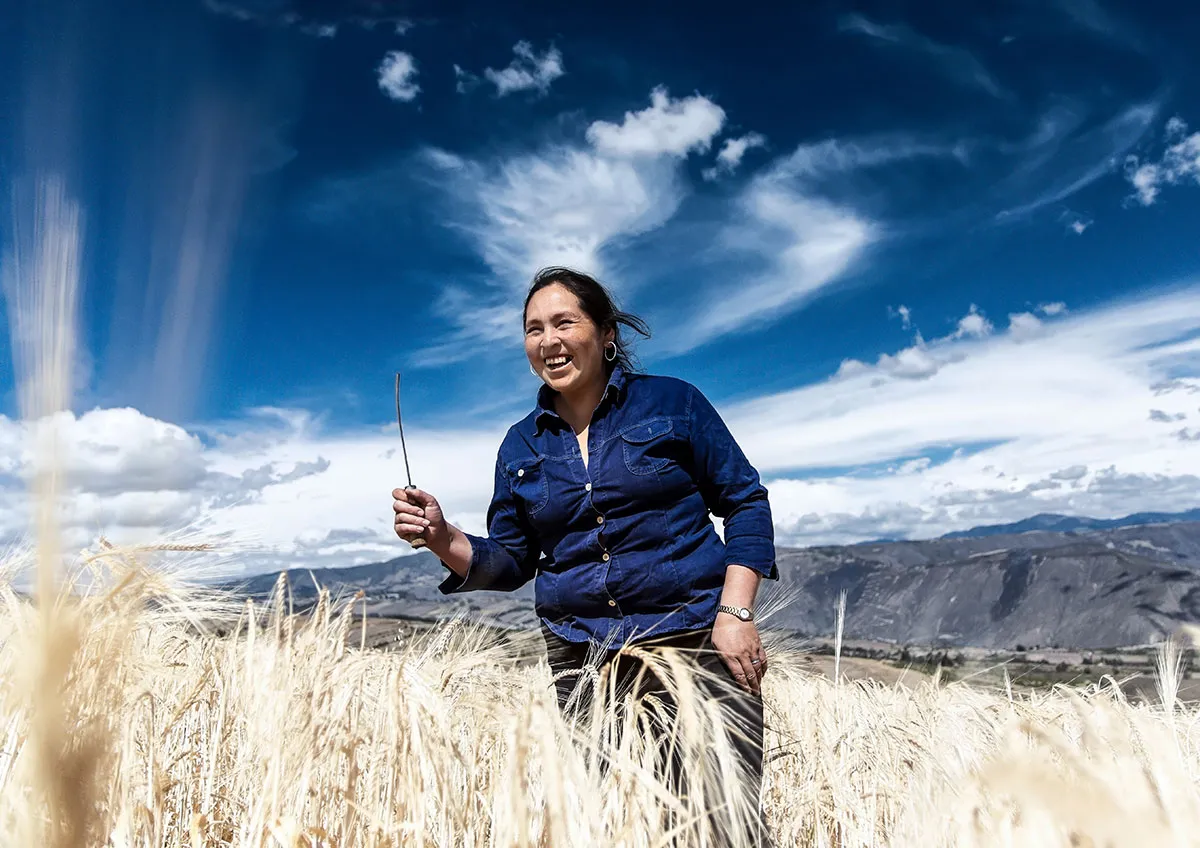 A woman smiles while walking through a lush field of wheat. Behind her is a bright blue sky with white clouds.