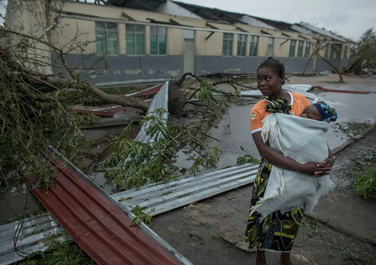 A woman holds her young child in a blanket while walking through a street filled with fallen tree branches and debris.