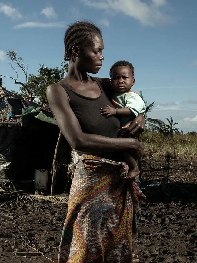 A woman holds her baby and looks at him. Behind her is a hut flanked by a couple of palm trees.