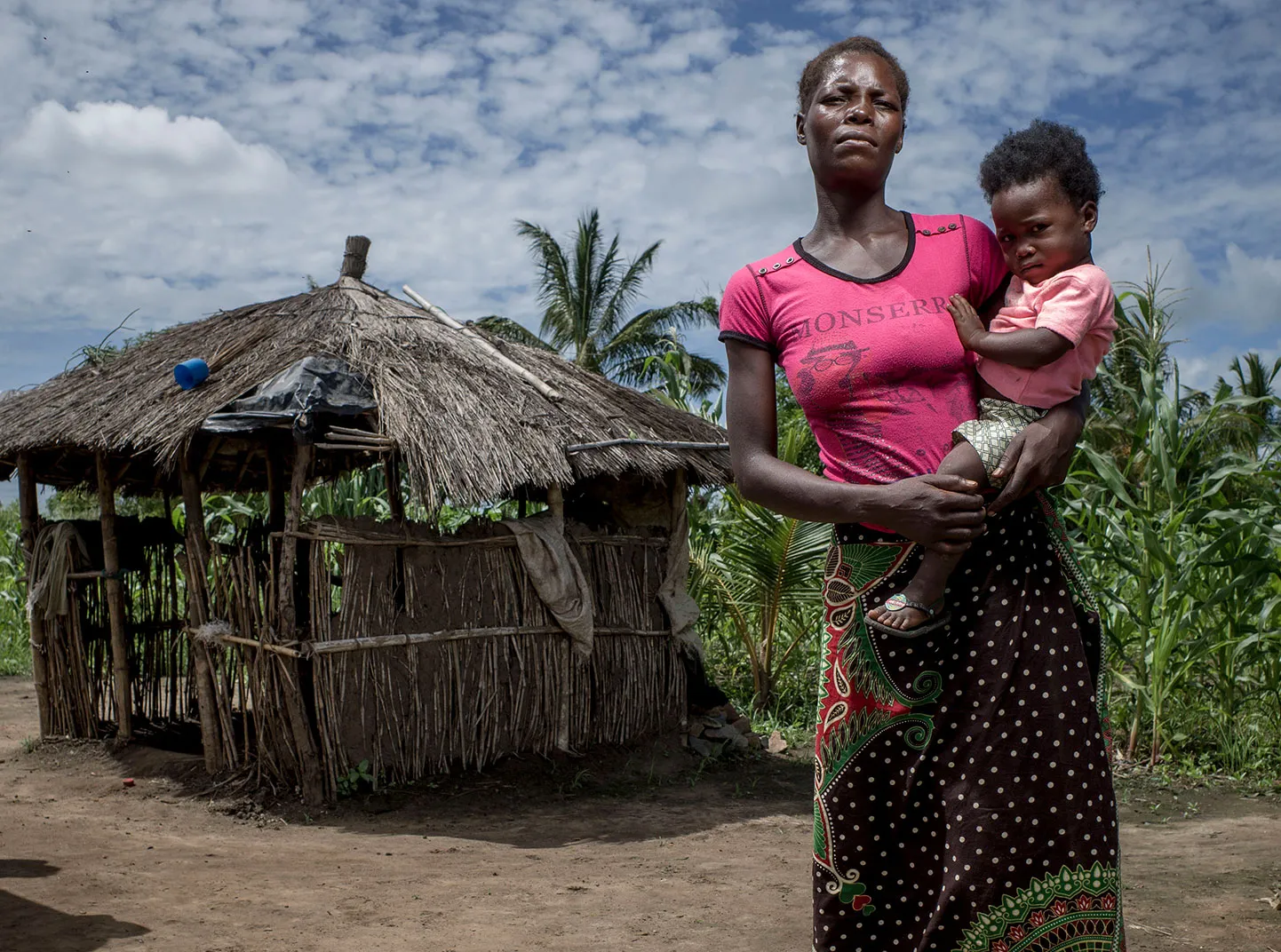 A woman holds her small child in front of a hut.