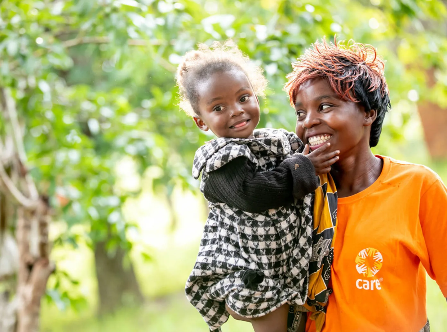 A CARE staffer wearing a bright orange CARE shirt smiles while holding a young girl wearing a black and white checked dress.