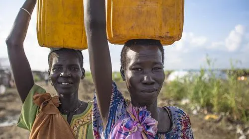 Two women pose for a picture with their newly-filled water containers on their head inside the Bentiu Protection of Civilian Area of the UN. There are about 50,000 people that have sought shelter and safety from the conflict in Sudan. CARE is committed to