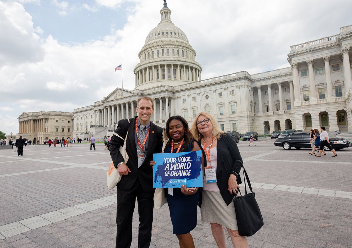 Three CARE Action advocates smile in front of Capitol Hill. One of them holds a sign that says, 