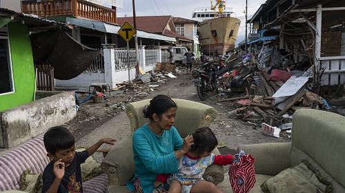 Dian, une survivante du tsunami, est assise avec ses enfants sur des canapés devant sa maison, avec un navire bloqué par le tsunami en arrière-plan.
