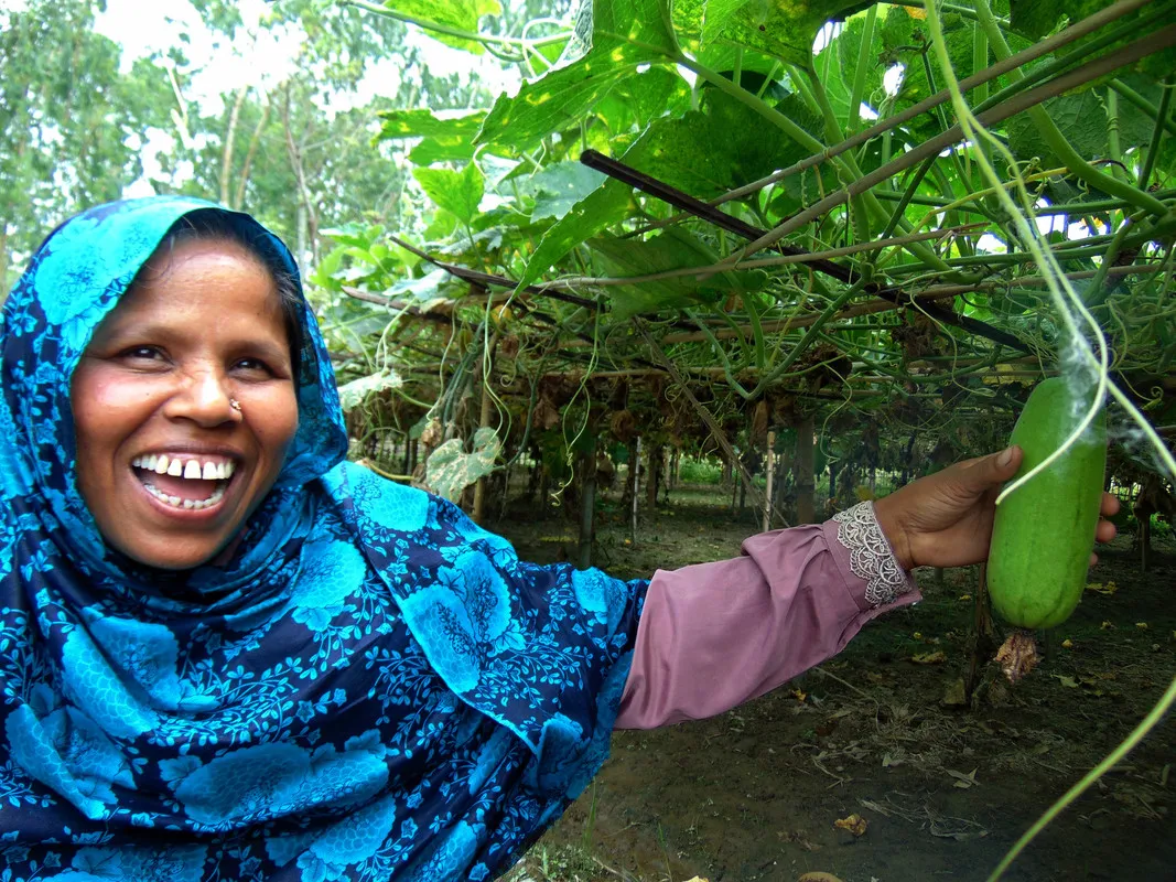 Uma mulher usando um lenço de cabeça azul brilhante abre um grande sorriso enquanto exibe algumas de suas colheitas.