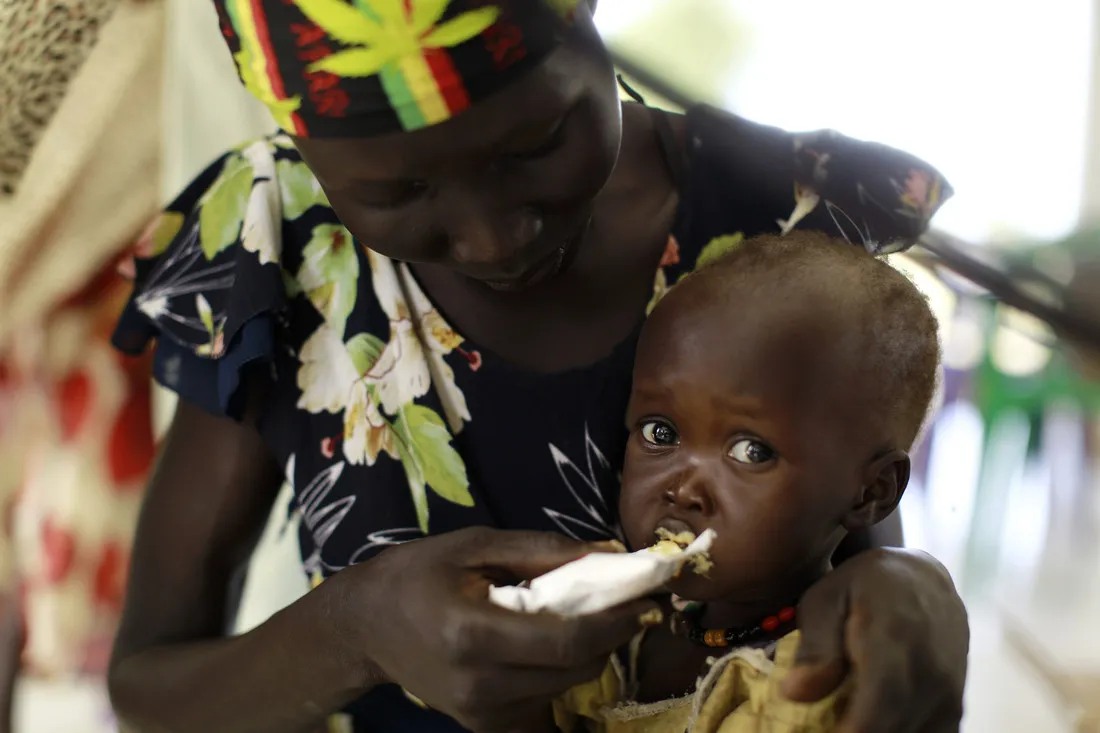 A woman hand-feeds a baby food. The baby is looking at the camera.