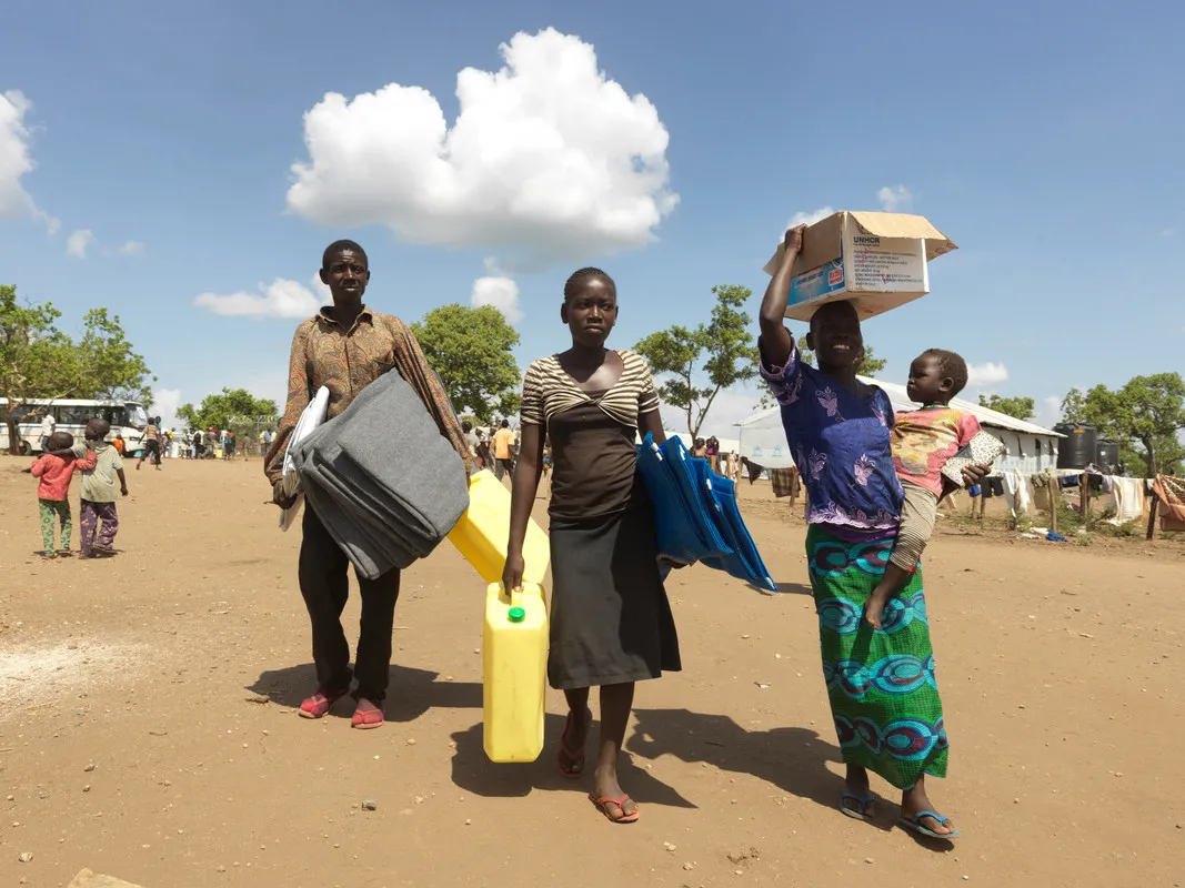 Two women and a man walk on a dirt road, all carrying blankets or buckets to hold water. The woman in front is balancing a cardboard box on her head with one arm, and carrying a baby with the other.