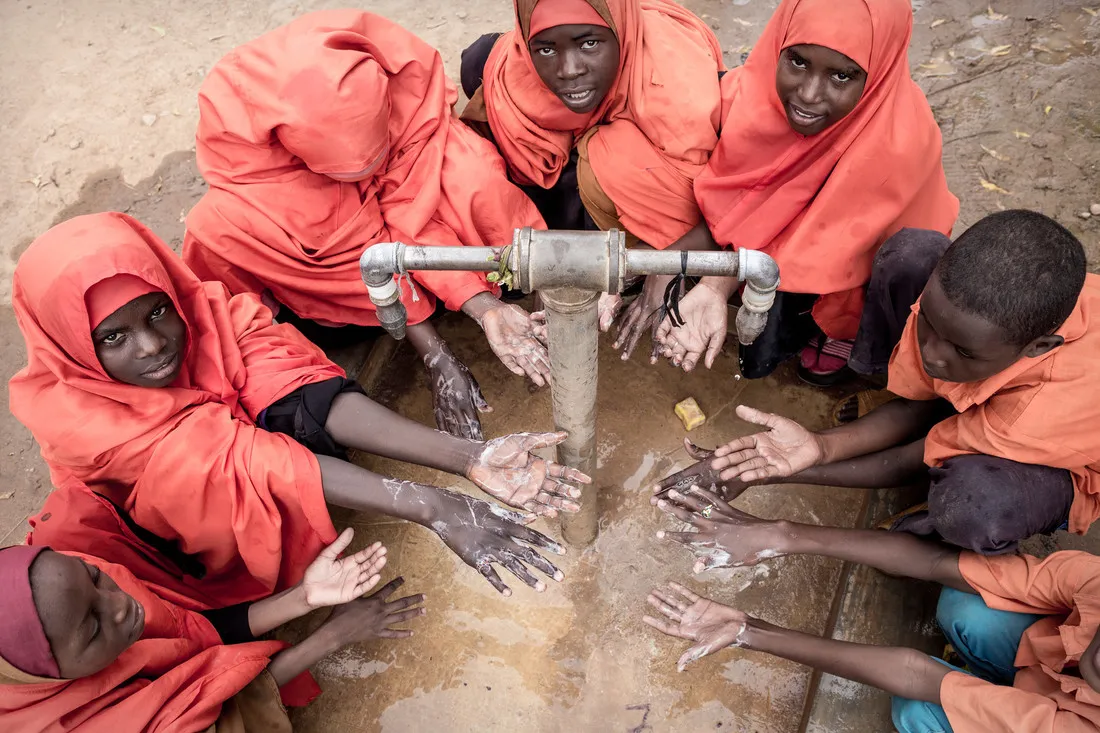 A group of kids wearing red garments hold out their hands underneath a spout of water. The device will help improve water sanitation and hygiene.