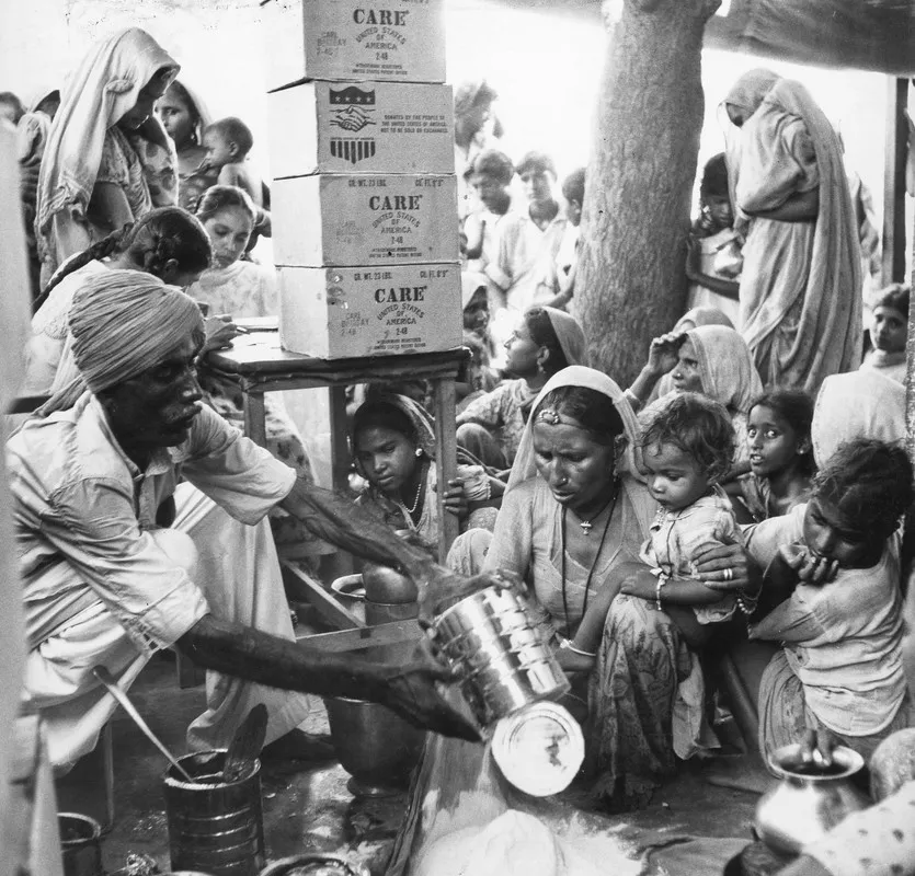 An Indian man wearing a headscarf pours flour out of a large can and into a bag. A mother and her young child watch. Behind them is a large group of men, women, and children sitting and standing. Four large CARE boxes are stacked on top of a nearby table.