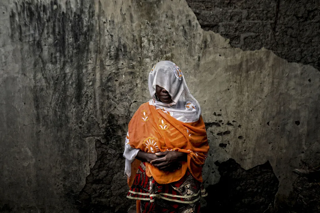 A woman stands against a wall with her hands folded in front of her. A white veil covers her face to protect her identity.