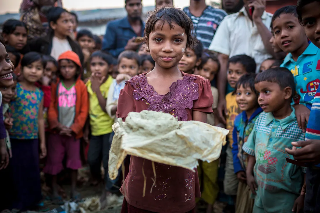 A young girl holds a cloth with a lump of clay up towards the camera. Behind her stand a large group of children.