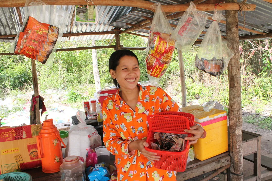 Uma mulher cambojana vestindo um robe estampado laranja brilhante sorri enquanto abre uma cesta vermelha cheia de contribuições em dinheiro para a VSLA.