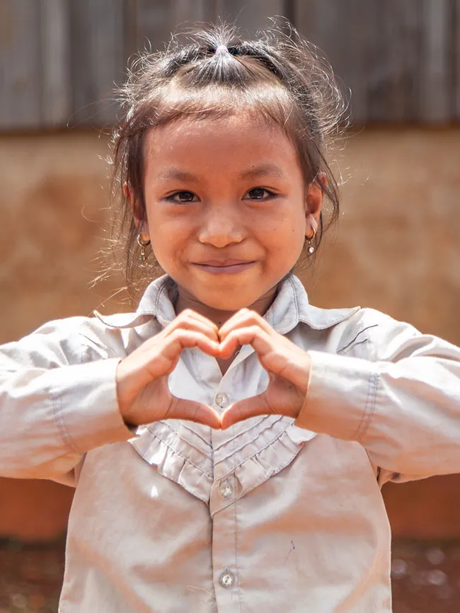 A young Cambodian girl smiles and makes a heart shape with her hands.