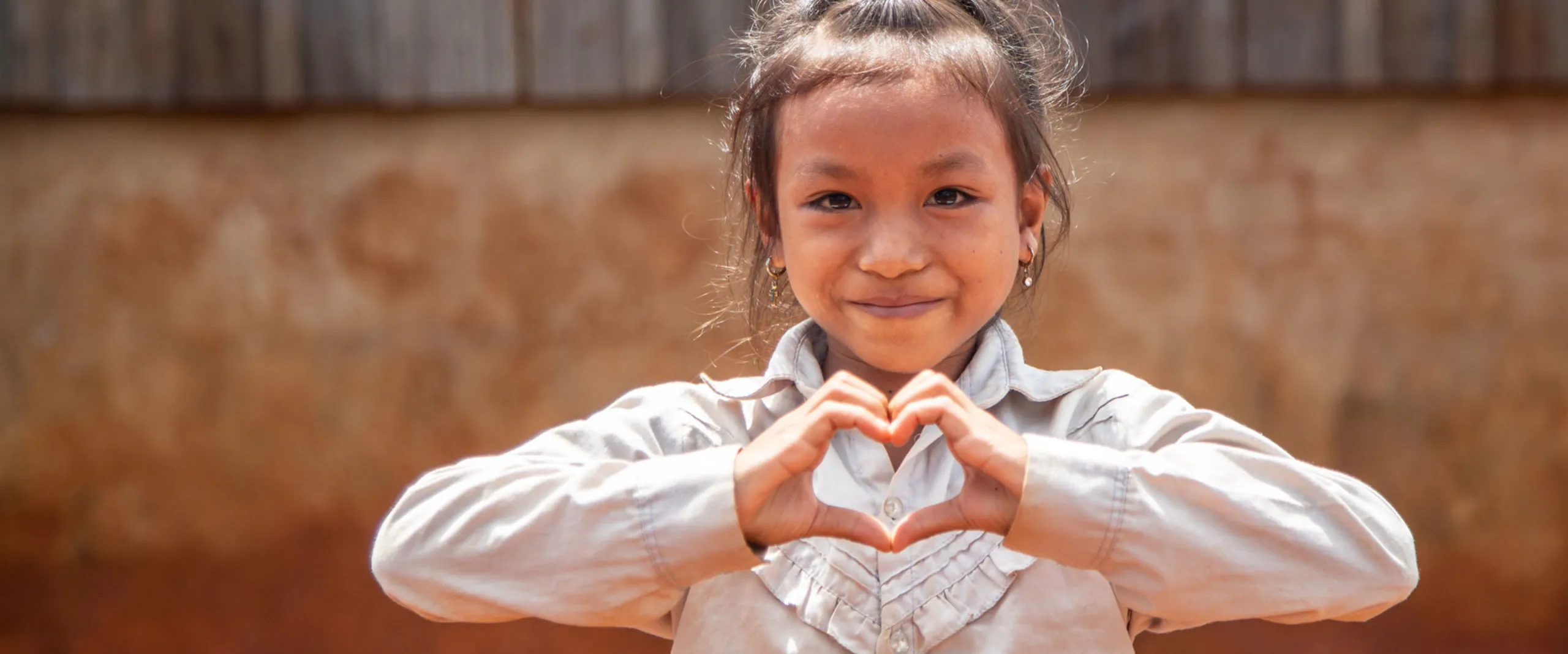A young Cambodian girl smiles and makes a heart shape with her hands.