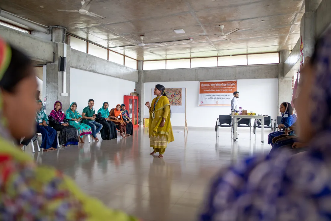 A woman wearing yellow walks around a room barefoot and speaks to a group of women.