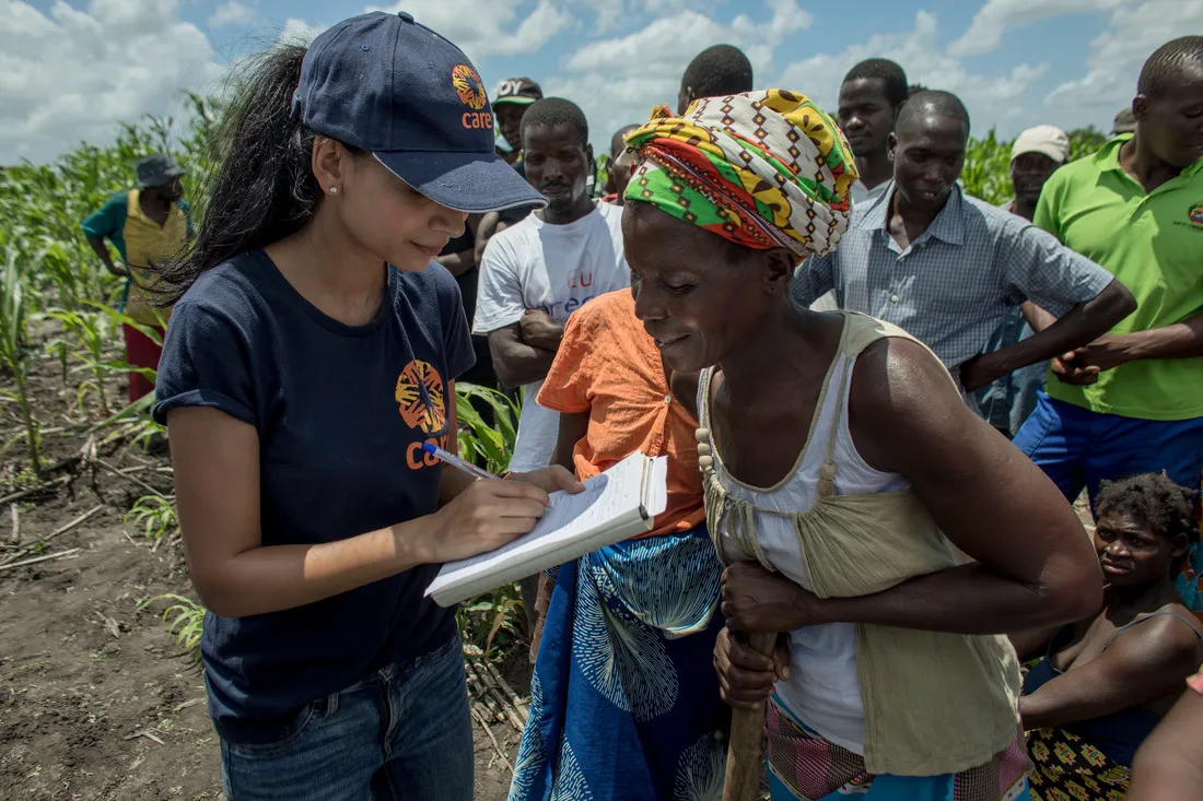 A female CARE staffer wearing a navy CARE shirt and hat jots down information in a notebook while speaking with a woman wearing a sleeveless shirt and a colorful headscarf.
