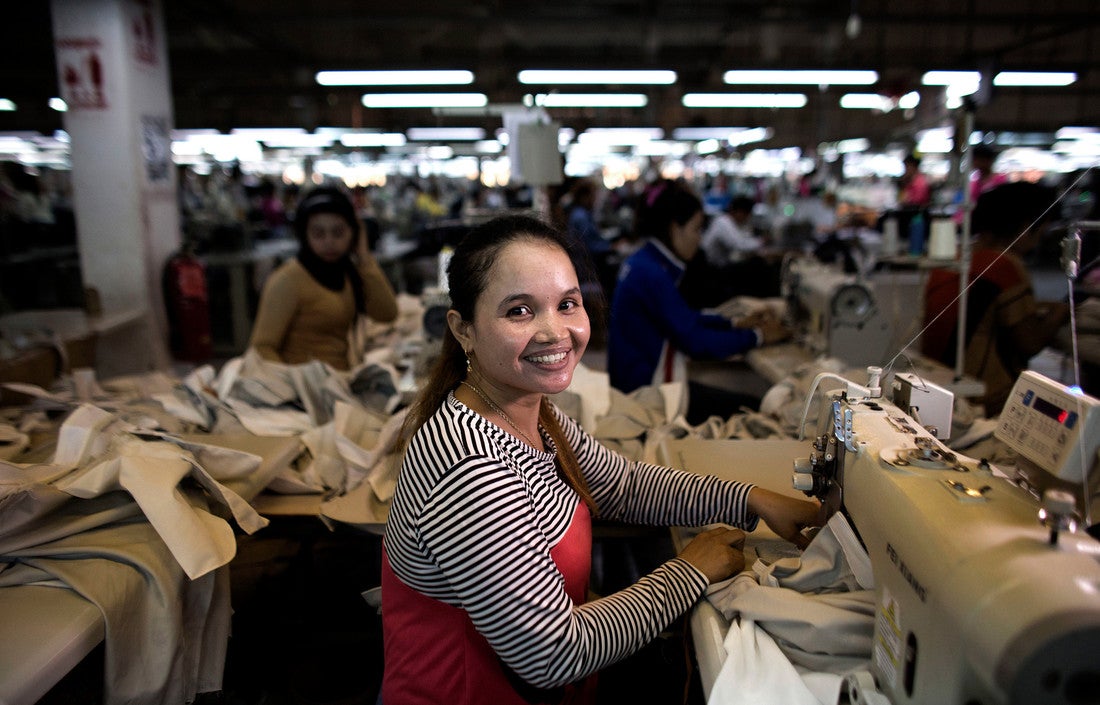 Uma mulher que trabalha em uma máquina de costura levanta os olhos de seu trabalho. Atrás dela, muitas outras mulheres trabalham em suas estações.