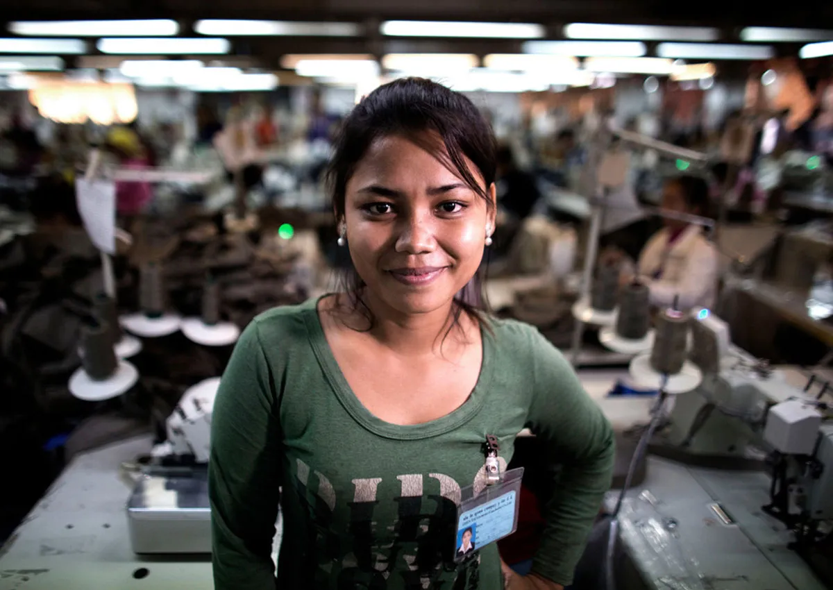 A Cambodian woman wearing a green long-sleeved shirt looks at the camera with a hand on her hip. Behind her, the interior of a garment factory is visible.