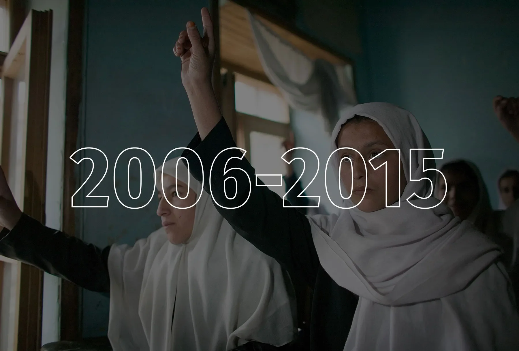 Two teenaged girls wearing dark long-sleeved shirts and white headscarves raise their right hands.