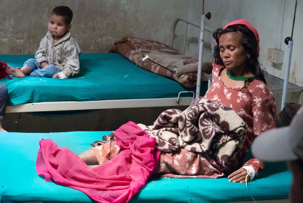 A woman and child sit in a small hospital room on two separate beds. Each is hooked up to an IV.