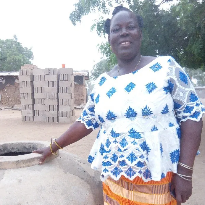 A woman in Ghana stands next to a stone workplace while in a dirt patch in from of cinder blocks.