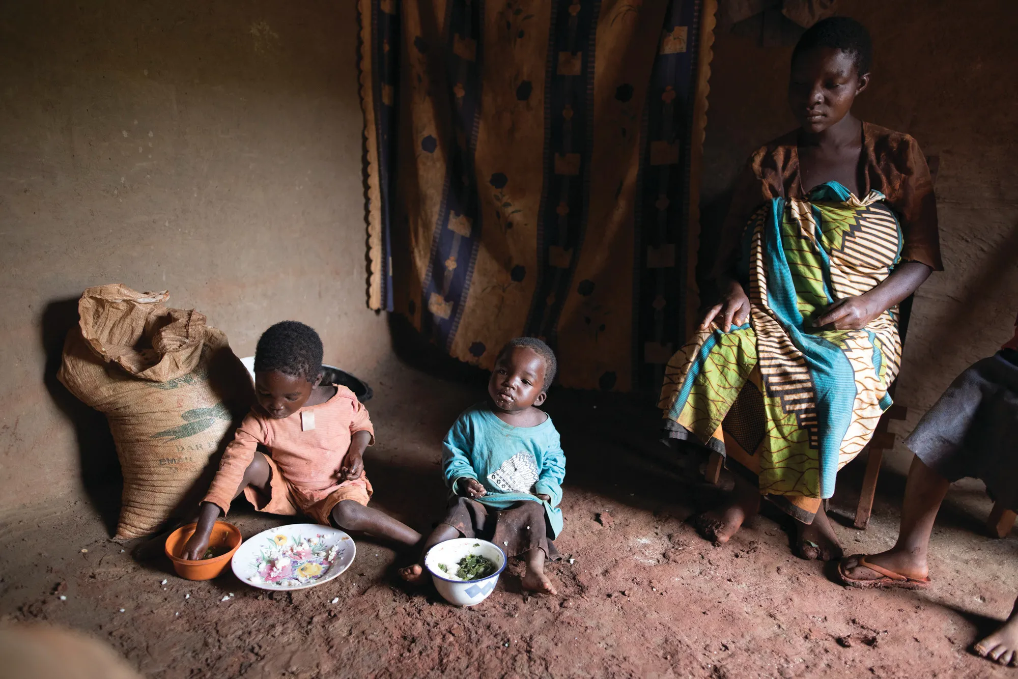 Une mère enceinte est assise sur une chaise en bois tandis que deux petits enfants avec des assiettes de nourriture devant eux sont assis sur le sol en terre battue à côté d'elle.
