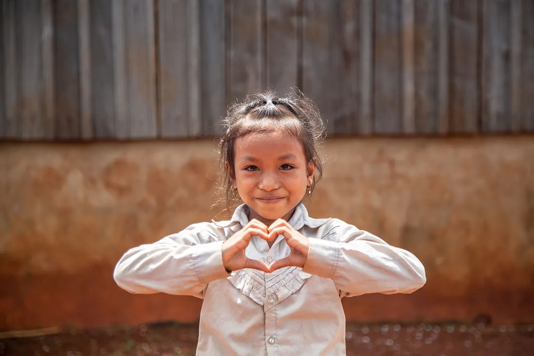 A young Cambodian girl smiles and makes a heart shape with her hands.
