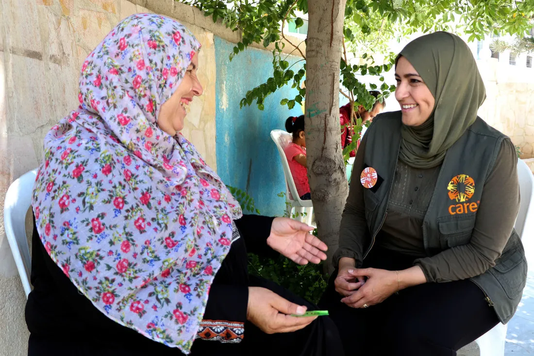 A woman wearing a green head scarf and a black CARE vest sits and speaks with a woman, who is smiling and gesturing with her hands.