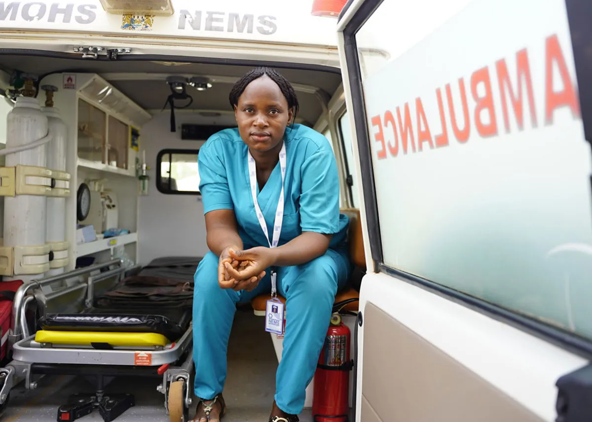 A woman wearing blue scrubs sits in the back of an ambulance.