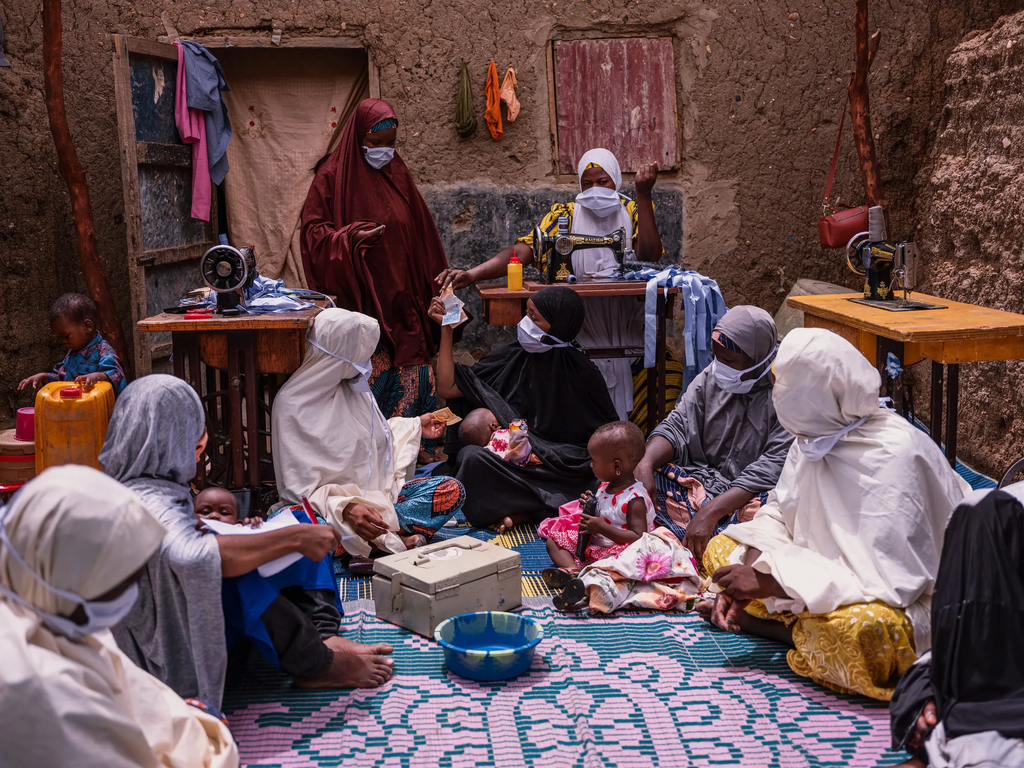 Um grupo de mulheres está sentado no chão em frente a várias mesas com máquinas de costura no Níger. Uma caixa de banco de metal está no centro do grupo.