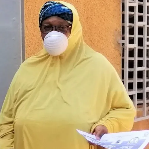 A woman in yellow with a mask on stands in front of a wooden door in Niger while holding papers.