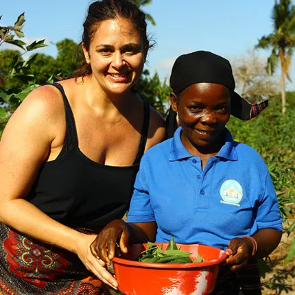 A woman wearing a tanktop poses with a kid. They're holding a bucket of green vegetables.