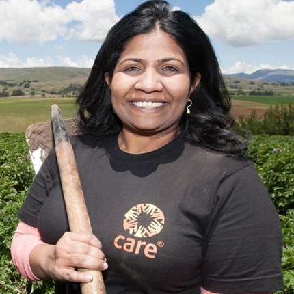 A woman wearing a black t-shirt with the CARE logo smiles while holding a garden hoe over her shoulder. She's standing in a large green field.