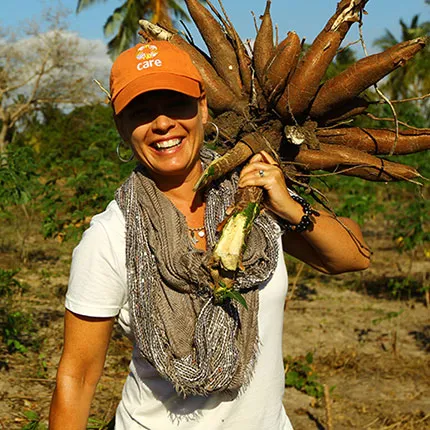 A woman wearing an orange CARE baseball cap smiles while holding a large bunch of veggies over her shoulder.