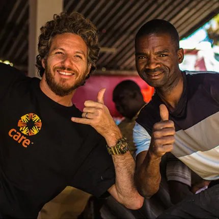 A man wearing a black CARE t-shirt poses next to a man wearing a striped polo shirt. Both are making friendly thumbs up gestures to the camera.