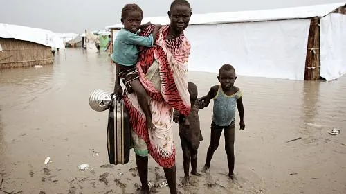 A woman carries a child on her shoulder and holds a suitcase. Next to her are two small children. They are walking through muddy dirt and there are white tents in the background.
