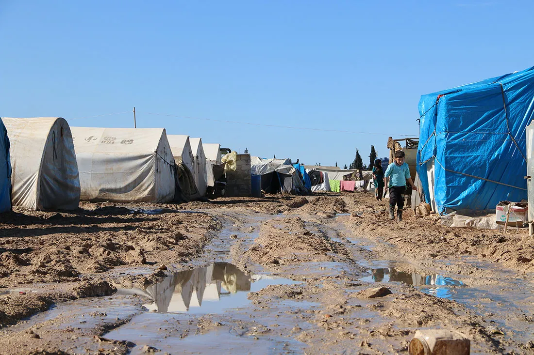 A landscape photo of a refugee camp in Syria. There are multiple large white tents lining a muddy street.