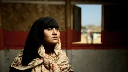 A woman wearing a black head scarf looks to the side while sitting in a house. Behind her, a small town is visible through an open window.