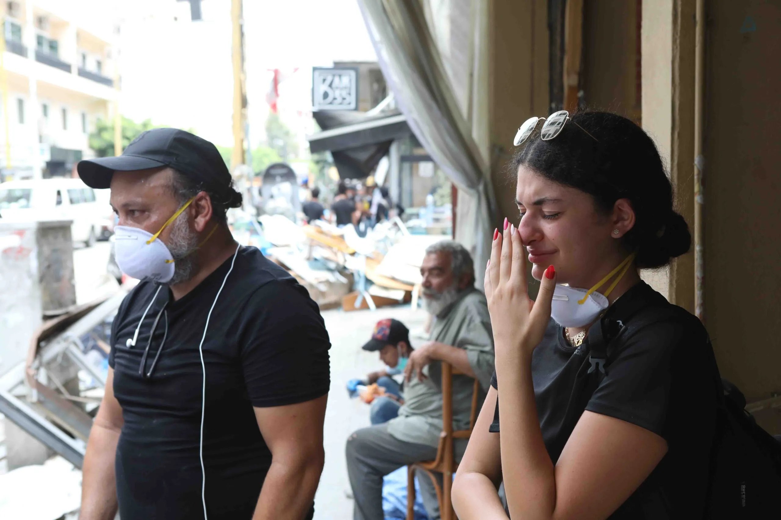 A man in a mask and a crying young woman with sunglasses on her head and a mask around her neck look at wrecked streets and buildings in Beirut.