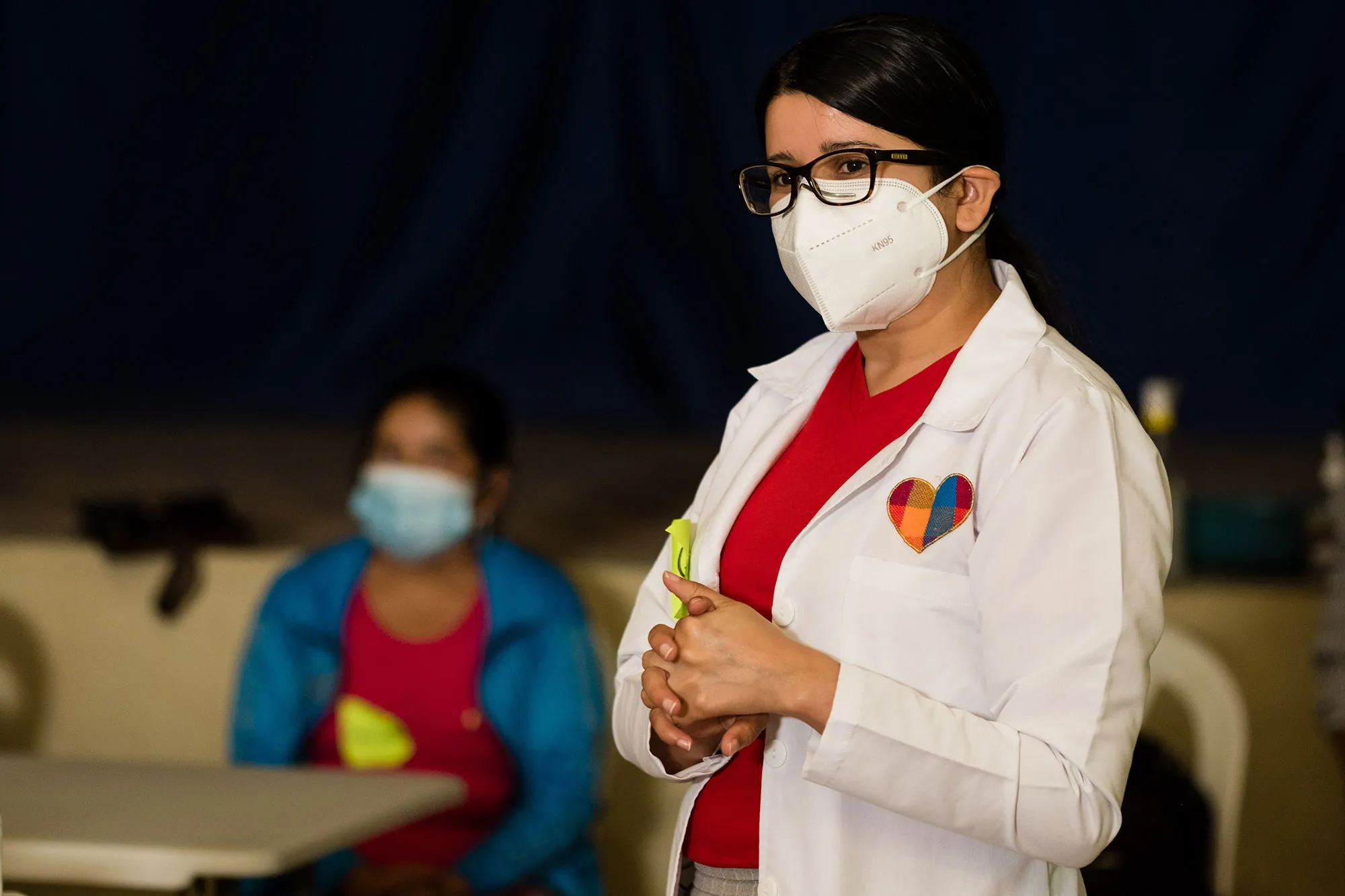 a woman in a lab coat and surgical mask stands.