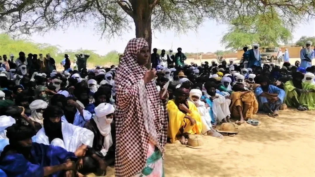 A woman in a checkered head wrap stands in front of a crowd of seated women.