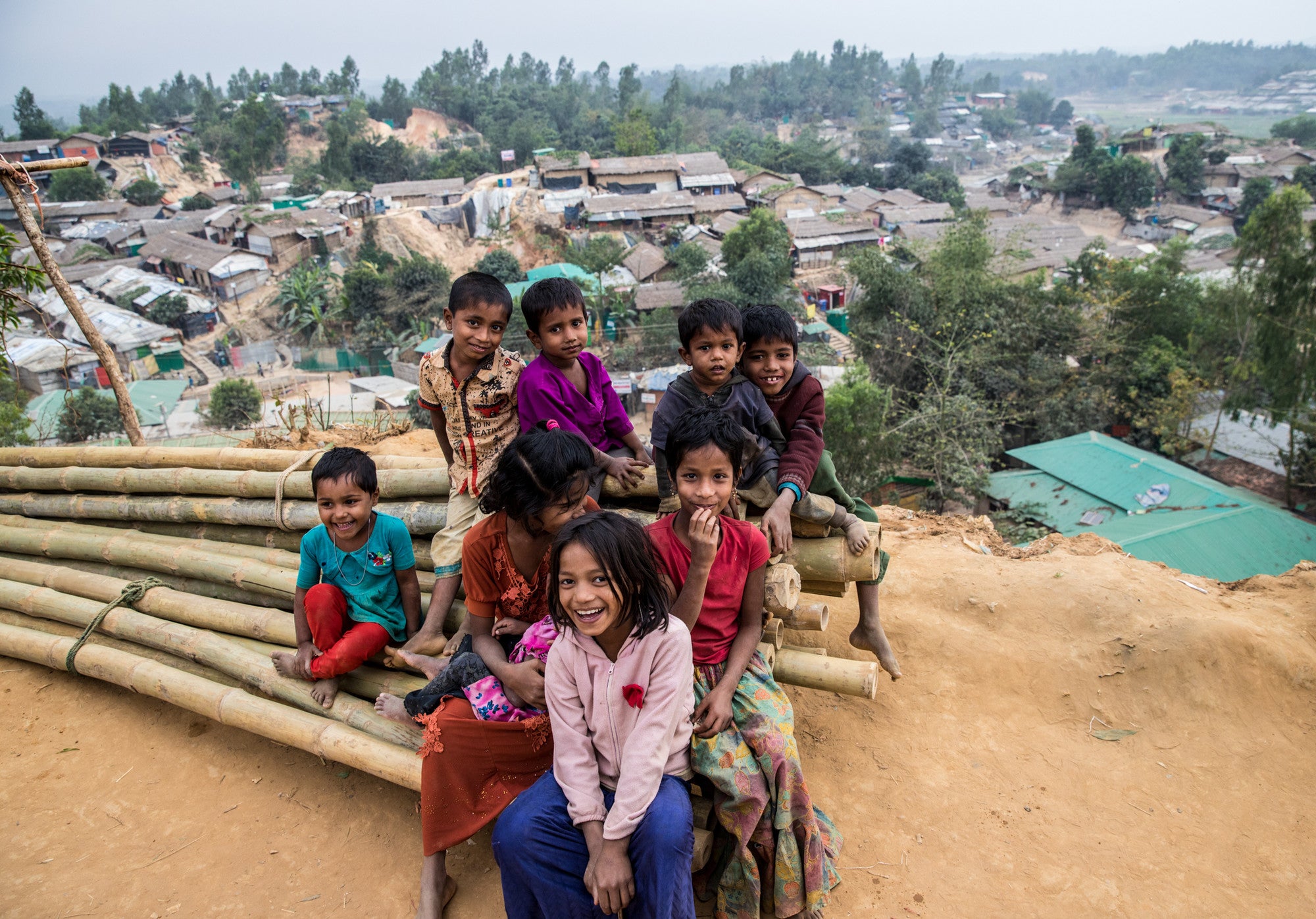 A group of children smile while sitting on a pile of large bamboo poles arranged in bundles.