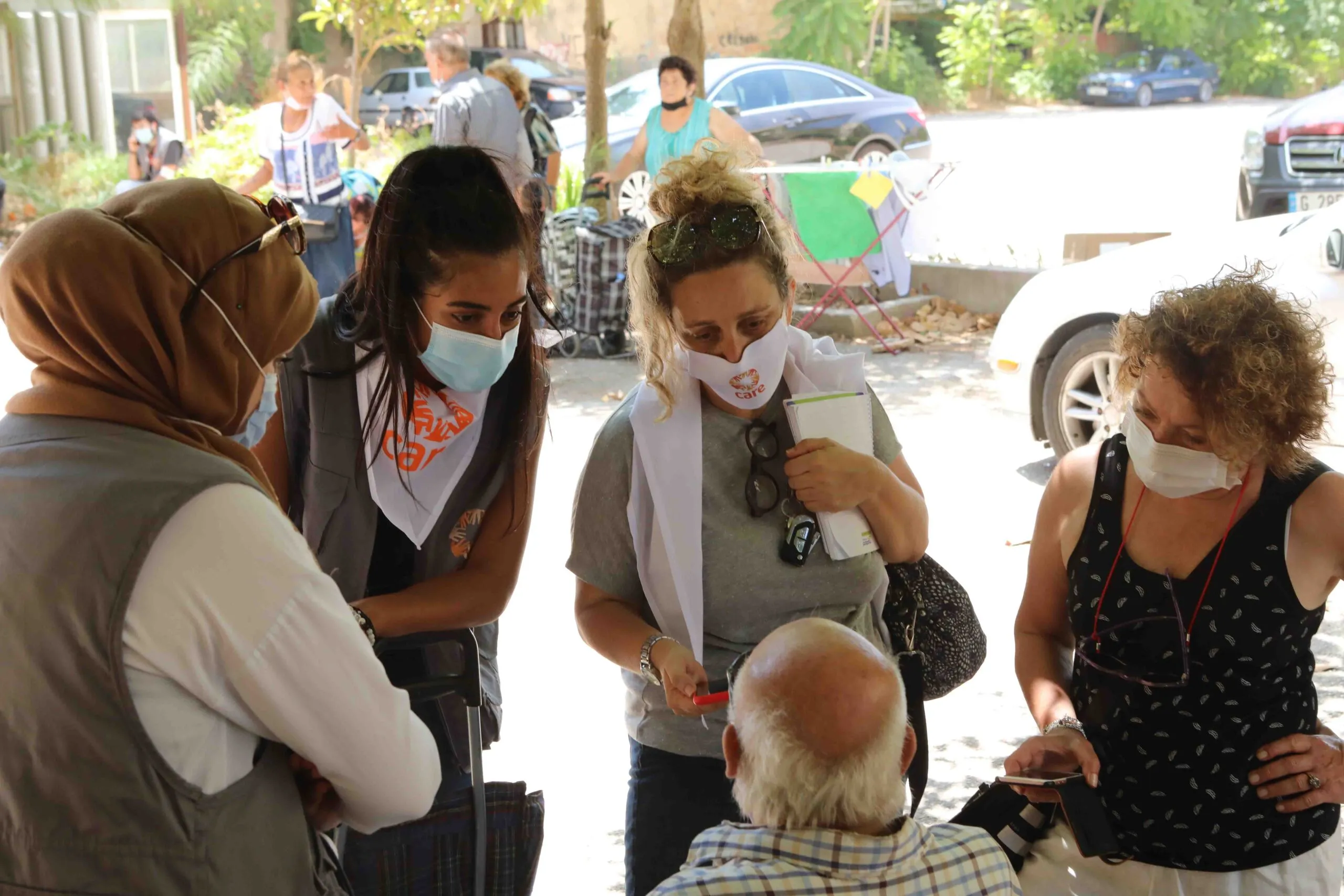 Four women in CARE face mask speak to a seated man.