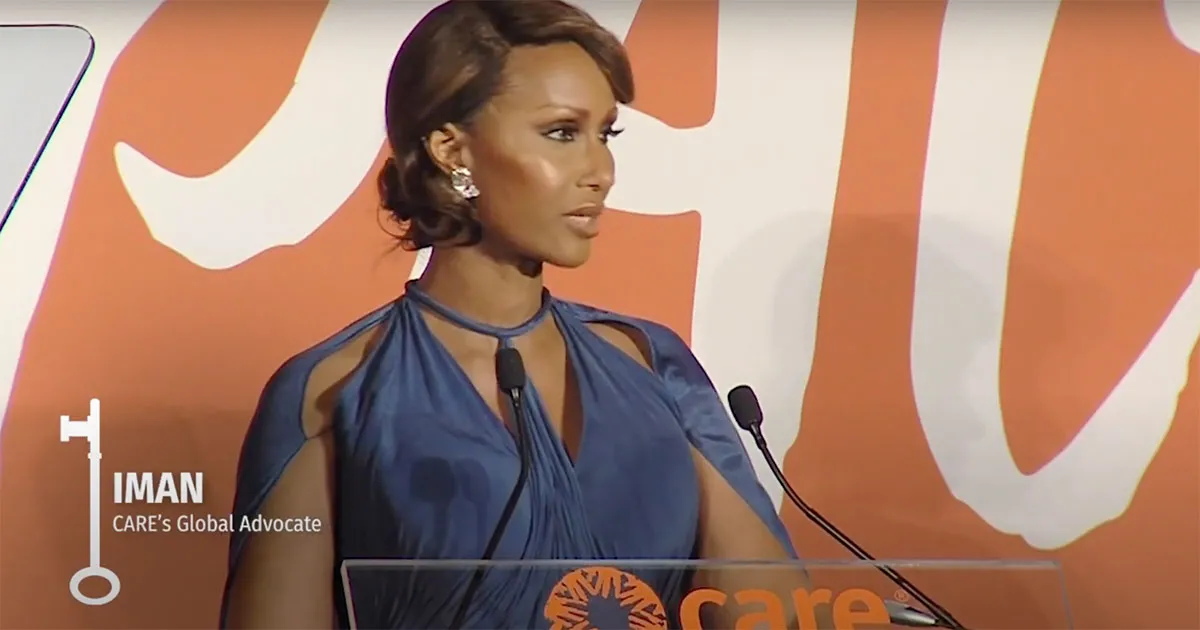 Iman wears a royal blue gown while accepting an award. She is speaking at a pedestal decorated with a CARE logo.