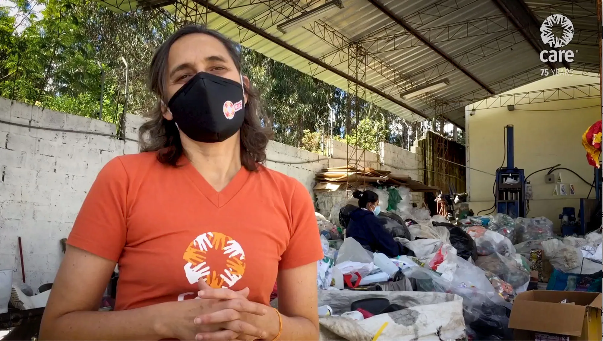 una mujer con una mascarilla se para frente a una planta de procesamiento de reciclaje mientras personas con máscaras trabajan detrás de ella.