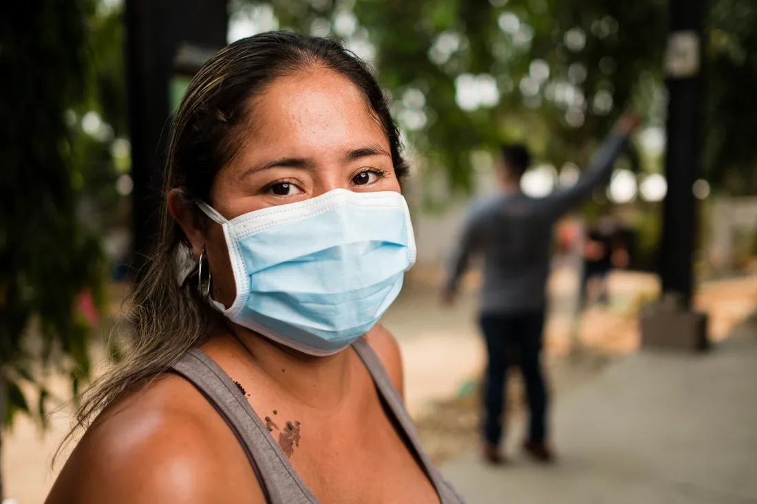A close-up of a Honduran woman with her body facing towards the right and her face looking straight at the camera. She is wearing a light brown tank top and her dark brown eyes are visible over her light blue medical face mask.