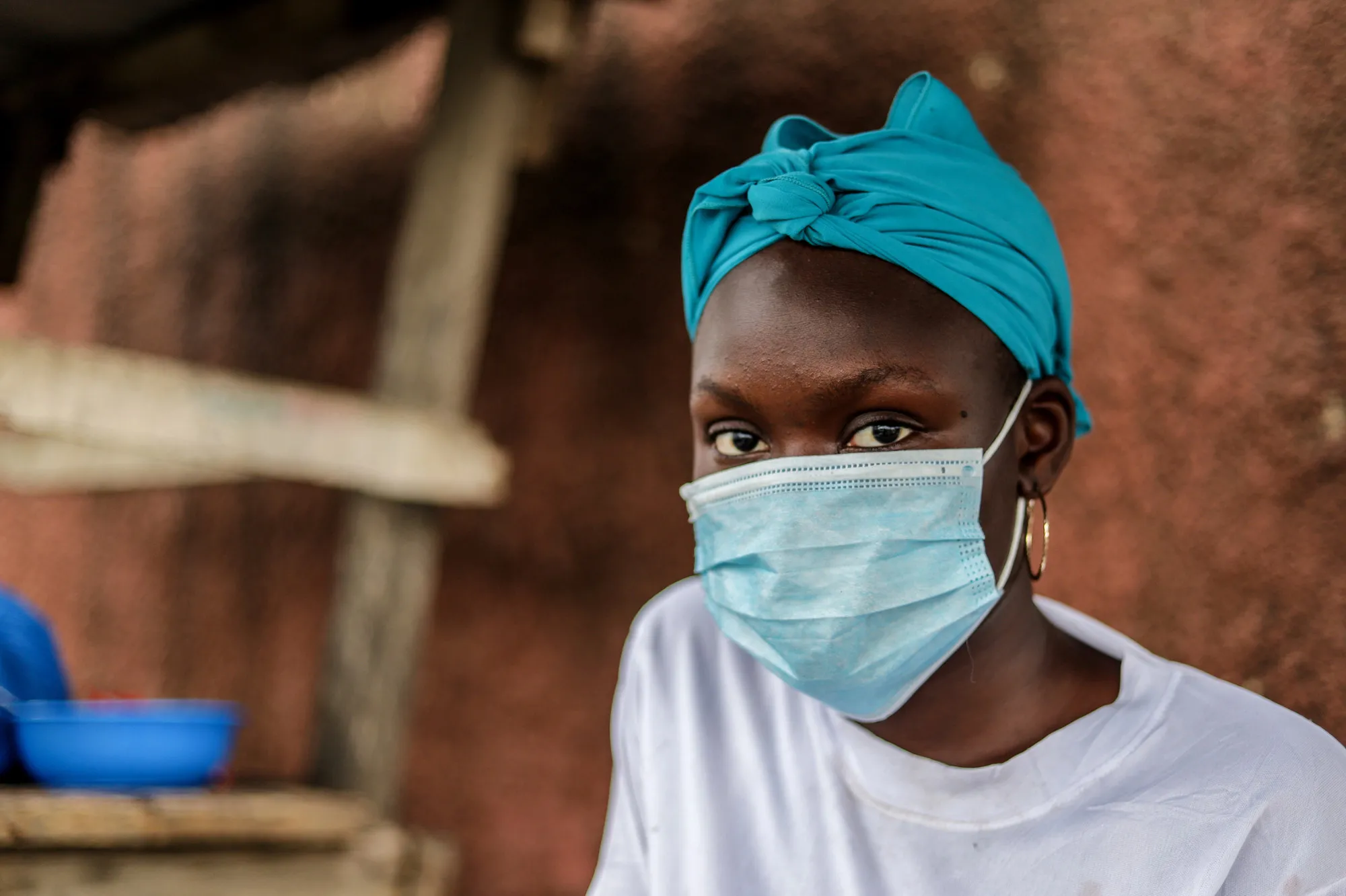 Woman in a head wrap wears a surgical mask in front of a dried mud wall