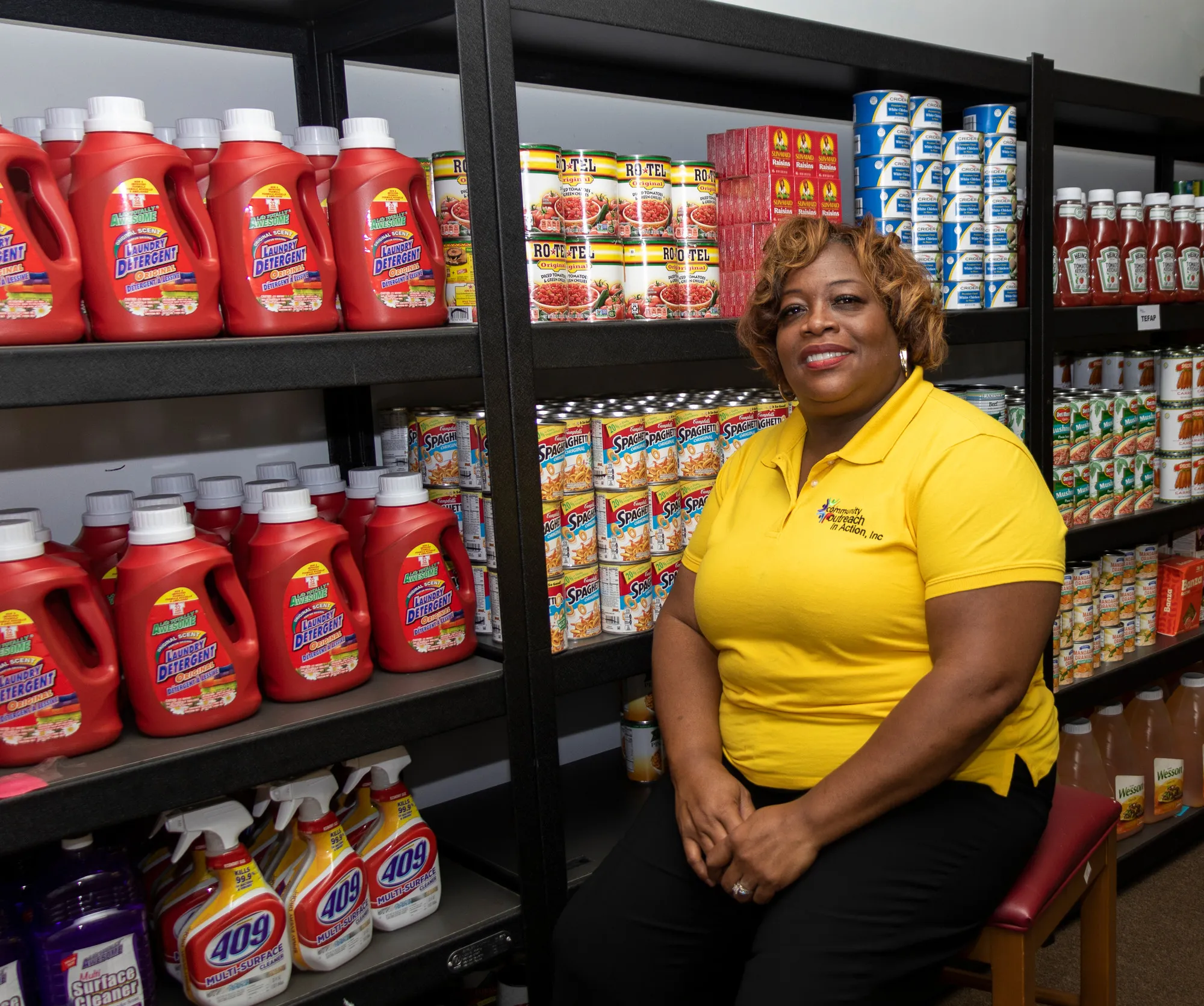 Una mujer administradora de un banco de alimentos se sienta frente a estantes llenos de alimentos.